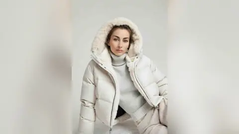 Young woman in a cozy white puffer jacket and turtleneck sweater posing against a neutral studio background.