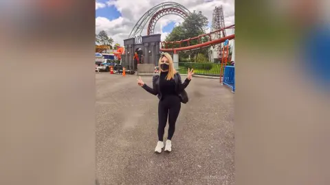 Happy woman in black outfit making peace signs at an amusement park with a large red roller coaster background.
