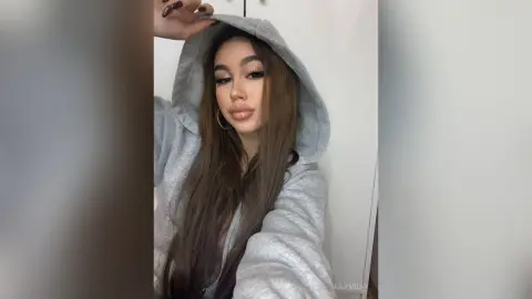 Young woman with long brown hair wearing a cozy grey hoodie in a casual indoor setting.