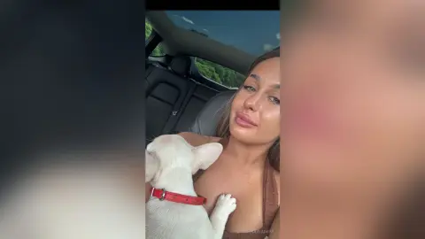 KaylaDepp poses in a car, wearing a brown top and looking at the camera with a white dog resting against her chest.