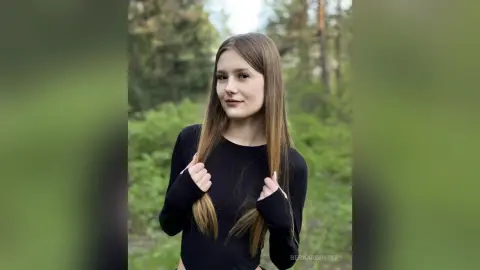 Young woman in a black long-sleeve shirt standing in a lush green forest with a serene expression.