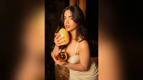 Elegant woman in a white slip dress holding golden decorative spheres in a warm, moody indoor setting.