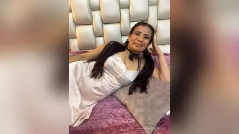 Smiling woman in a white satin dress and black choker relaxing on a bed with a quilted headboard.