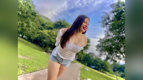 Smiling young woman in a white off-the-shoulder top and denim shorts enjoying a sunny day in a lush green park.