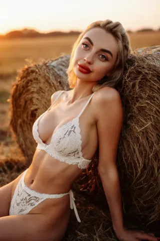 Blonde woman in white lace lingerie leaning against hay bales in a golden sunset field.
