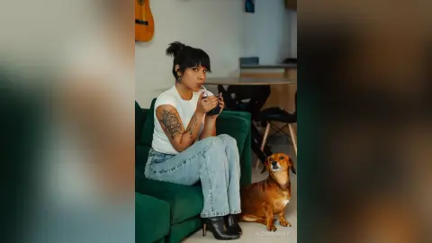 Tattooed woman in white t-shirt and jeans sitting on a green sofa with a brown dog in a cozy indoor setting.