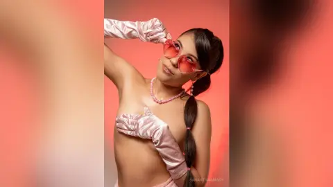 SamanthaHarvey poses against a red background wearing pink sunglasses, a pink beaded necklace, and long pink satin gloves.