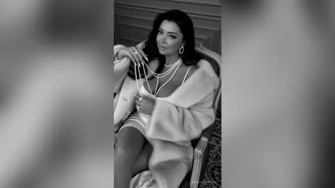 Elegant woman in a white fur coat and pearl necklace sitting glamorously in a vintage indoor setting.