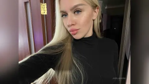 JudiJadore poses in a black long-sleeved turtleneck, looking directly at the camera with her long blonde hair down.