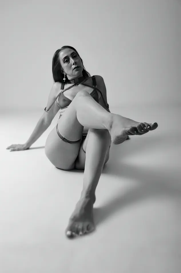 PrettyLady74 — Elegant woman in minimalist lingerie posing gracefully on a studio floor in a moody black and white portrait.