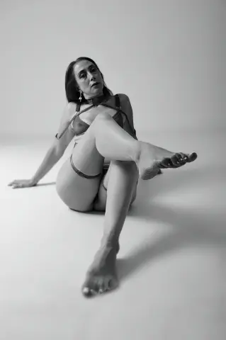 Elegant woman in minimalist lingerie posing gracefully on a studio floor in a moody black and white portrait.
