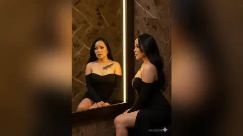 Elegant woman in a black off-the-shoulder dress looking into a marble mirror in a moody, luxury setting.