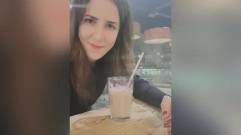 Smiling young woman in a black sweater enjoying a creamy milkshake at a cozy cafe at night.