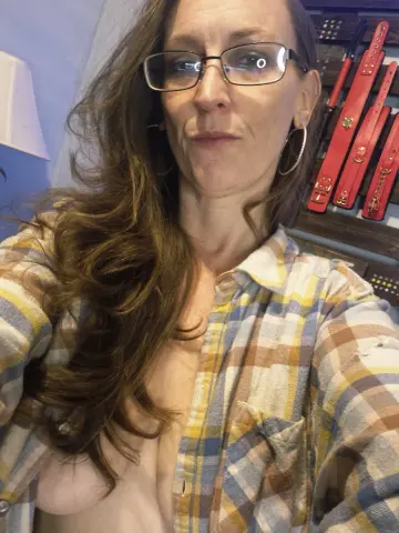 Woman with long brown hair and glasses wearing an open yellow plaid shirt in a casual indoor setting.