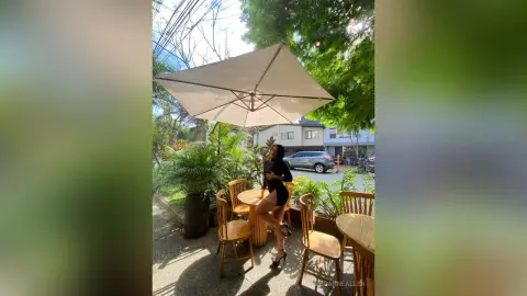 Stylish woman in a black romper sitting at an outdoor cafe table under a large patio umbrella on a sunny day.