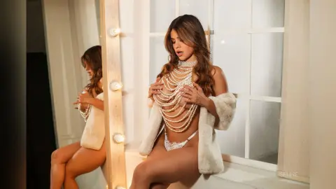 Glamorous woman in white lace lingerie and pearl necklace sitting by a bright window in a soft, elegant mood.