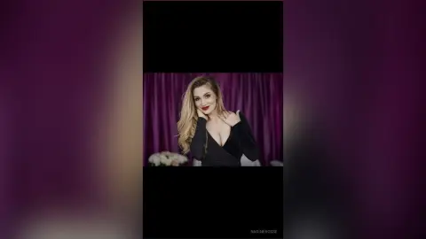 Elegant woman in a black velvet dress with red lipstick posing against purple curtains in a moody setting.
