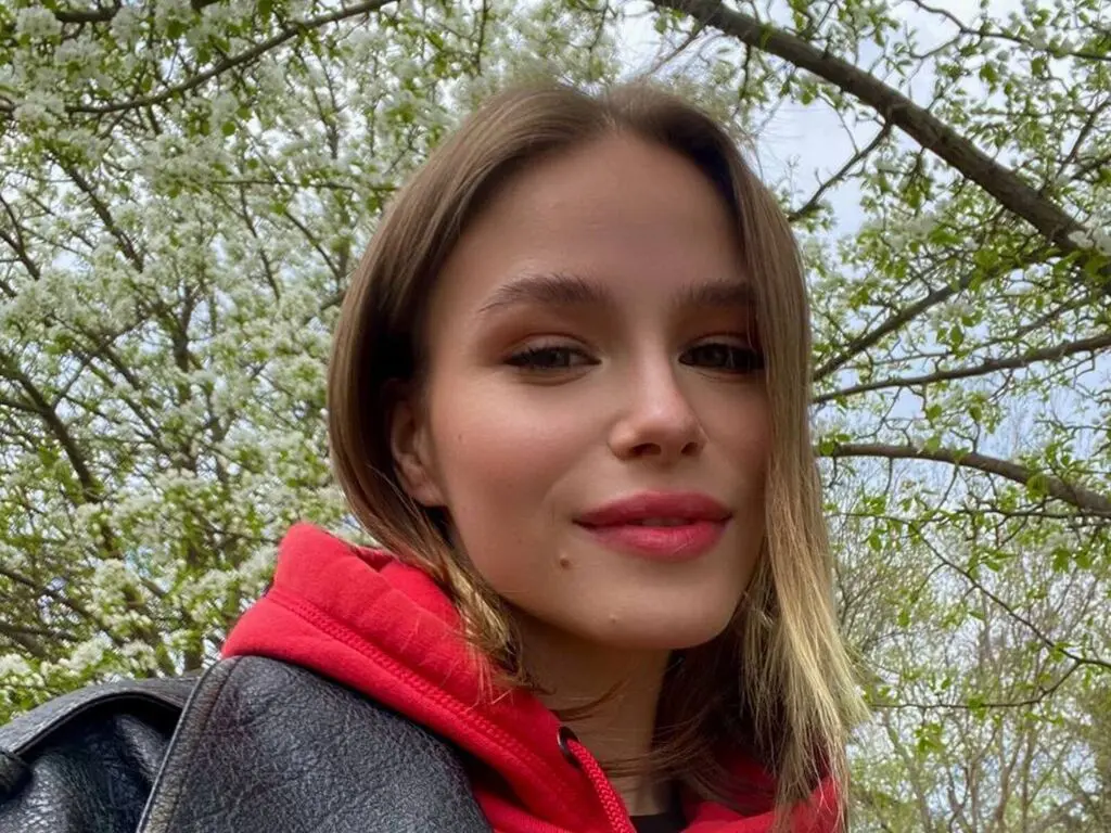 Smiling young woman in a red hoodie and black leather jacket standing under blooming white spring trees.