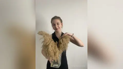 Smiling woman in a black sleeveless top holding a large bouquet of dried pampas grass in a bright room.