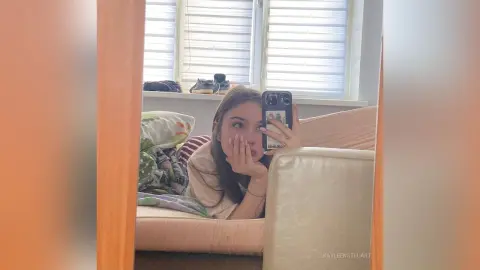 Young woman in a casual beige top taking a cozy mirror selfie on a living room sofa.