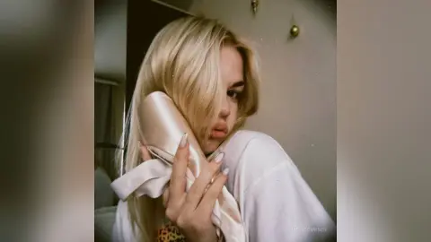Blonde woman in a white shirt holding a beige satin fabric in a soft, moody indoor setting.