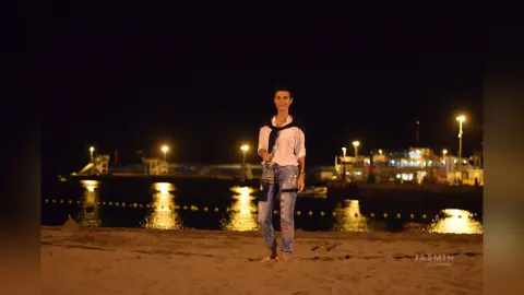 Smiling woman in a white shirt and denim jeans standing on a beach at night under glowing pier lights.