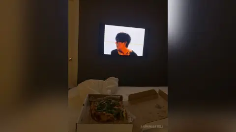 A pizza box sits on a bed in a dimly lit room with a movie playing on the TV screen.