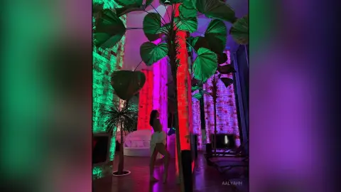 Aalyahh stands naked in a room with large tropical leaves and red and green lighting, showing her bare back and legs.