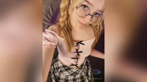 Blonde woman in glasses and a pink corset top with a plaid skirt posing in a soft, dreamy indoor setting.