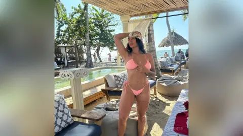 Young woman in pink bikini relaxing at a tropical beach resort under a thatched sunshade.