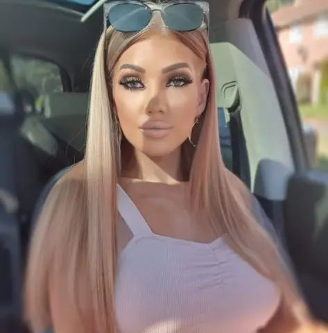 BritishTastyCherryxo with long blonde hair wearing a white tank top sits in a car.