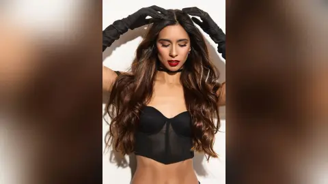 Glamorous woman with long wavy hair wearing a black corset top and red lipstick against a white background.
