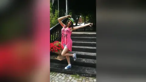 Cheerful young woman in a pink graphic tank top and denim shorts posing on outdoor wooden stairs.