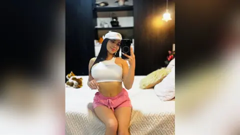 Young woman in white crop top and pink shorts relaxing on a bed in a cozy, dimly lit bedroom.