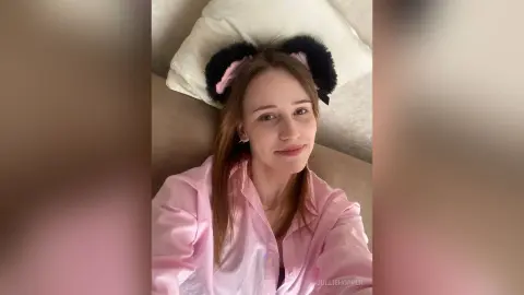 Smiling young woman in a pink button-down shirt wearing black fuzzy headband ears while relaxing on a couch.