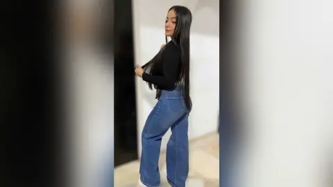 Young woman with long dark hair wearing a black long-sleeve top and blue wide-leg jeans in a minimalist setting.