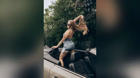AdelinaKael poses in a silver dress while sitting in a convertible car, running her hands through her long blonde hair against a backdrop of green trees.