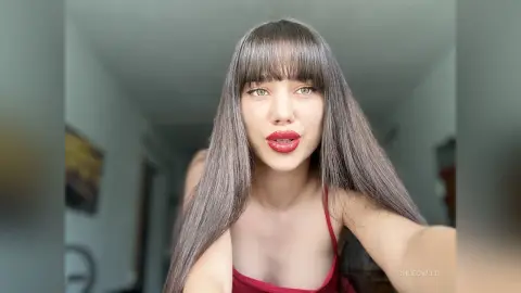 Young woman with long straight hair and red lipstick wearing a red tank top in a bright indoor setting.