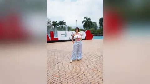 Woman in white crop top and light blue wide-leg pants standing by a pool near large red letters.