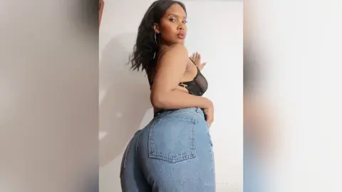 Confident woman in a black sheer bralette and blue denim jeans posing against a white wall.