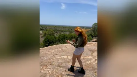 Redheaded woman in a green shirt and striped shorts standing on a rocky cliff overlooking a scenic forest view.