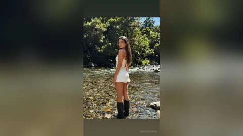 Young woman in a white romper and black boots standing in a shallow rocky river surrounded by lush green trees.