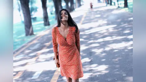 Smiling woman in a red polka dot summer dress walking along a sunny park path.