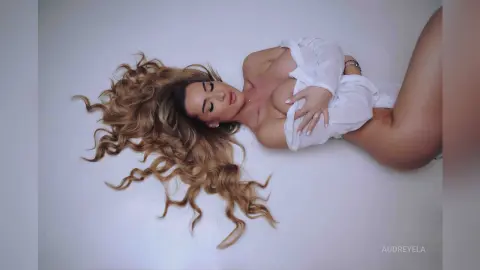 Glamorous woman with long wavy blonde hair wearing a white shirt lying on a white floor in a dreamy mood.