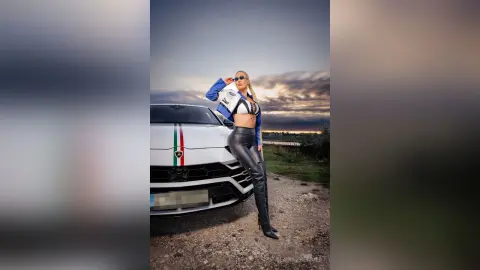 Stylish woman in a blue blazer and black leather pants posing confidently next to a white sports car at sunset.