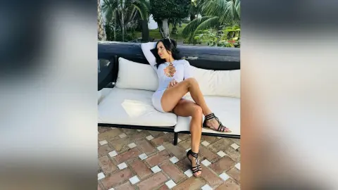 Stylish woman in a white cutout bodysuit relaxing on a white outdoor lounge sofa in a tropical garden setting.