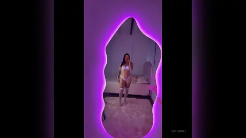 NatiSweet poses in a white bra and panties set, standing in front of a purple-lit mirror in her bedroom.