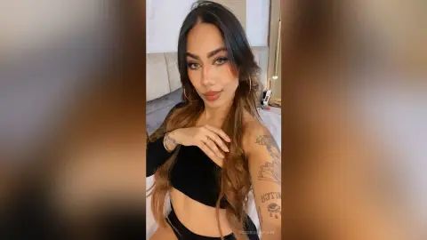 VictoriaFontaine poses in a black crop top, showing off her long brown hair and arm tattoos in a bedroom setting.