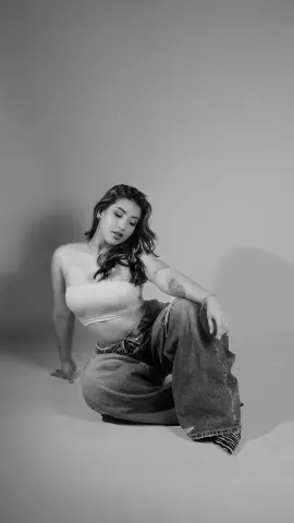 Moody black and white portrait of a young woman in a white tube top and baggy denim jeans sitting on a studio floor.