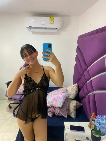 Smiling woman in a black lace bodysuit taking a mirror selfie in a cozy bedroom setting.